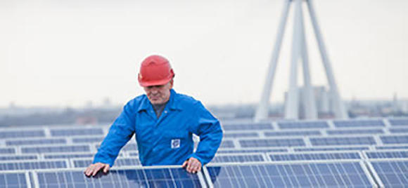 A man wearing an orange helmet and blue service suit inspects solar panels on a rooftop.
