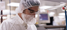 A man in half-profile wearing laboratory safety protection looks downward at an object off-camera. 
