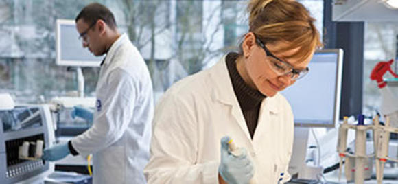 A woman wearing lab safety equipment uses a pipette in the lab, her coworker visible in the background.